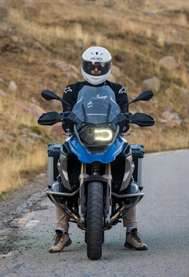 A person wearing a white motorcycle helmet and black jacket sits on a blue adventure motorcycle on a rural road. The background shows dry grass and rocky terrain.