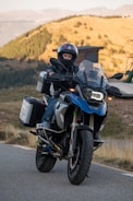 A person wearing a helmet is riding a large touring motorcycle on a paved road. The bike is equipped with saddle bags and has a rugged appearance, indicating it's suitable for long-distance travel. The background shows a scenic landscape with rolling hills and sparse vegetation under a clear sky.
