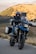 A person wearing a helmet is riding a large touring motorcycle on a paved road. The bike is equipped with saddle bags and has a rugged appearance, indicating it's suitable for long-distance travel. The background shows a scenic landscape with rolling hills and sparse vegetation under a clear sky.