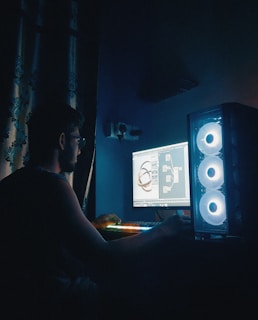 A person is working on a computer in a dimly lit room. The setup includes a desktop PC with a transparent case and illuminated cooling fans. The display shows a complex 3D modeling software with intricate graphics. The room has dark curtains and the ambient lighting is primarily from the computer and keyboard, creating an atmospheric glow.