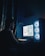 A person is working on a computer in a dimly lit room. The setup includes a desktop PC with a transparent case and illuminated cooling fans. The display shows a complex 3D modeling software with intricate graphics. The room has dark curtains and the ambient lighting is primarily from the computer and keyboard, creating an atmospheric glow.