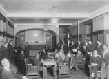 A group of men in suits gather in a room with wooden chairs and bookshelves. The room features a high ceiling with hanging lights and a brick fireplace at the far end. Some men are seated, while others stand around the perimeter, engaging with one another.
