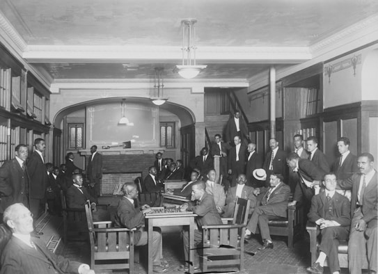A group of men in suits gather in a room with wooden chairs and bookshelves. The room features a high ceiling with hanging lights and a brick fireplace at the far end. Some men are seated, while others stand around the perimeter, engaging with one another.