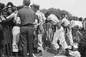 A group of uniformed officers, including individuals with armbands, are interacting with a crowd of people behind a metal fence. Some officers appear to be speaking or gesturing towards people, while one is pushing against the fence. Behind them, numerous people are observing the situation, holding signs. The scene appears to be tense.