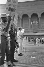 A group of people is gathered in front of a large building holding signs with messages related to civil rights and freedom. They appear to be engaged in a protest or demonstration. The architecture of the building features arches and columns, and several flags are visible in the background. The people in the foreground are diverse in terms of age and attire, indicating a mixed group participating in the event.
