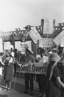 A group of people are gathered in a protest or rally, holding various signs with messages advocating for voting rights, democracy, and representation. Some of the signs read 'Fighting for the right to vote,' 'Freedom now,' 'We shall overcome,' and '1 million Mississippi Negroes deserve representation.' The crowd appears engaged and focused on the cause.