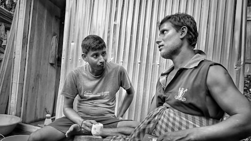 Two men are sitting and engaging in conversation in what appears to be an informal outdoor setting. One man holds a cup while the other listens attentively, with both wearing casual clothing. The background consists of corrugated metal and wooden elements, suggesting a rustic environment.