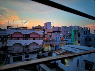 Urban setting with multiple residential buildings at sunrise or sunset, showcasing a mix of architectural styles. The sky has a gradient of blue and orange hues. There are utility lines in the foreground and a billboard in the background.