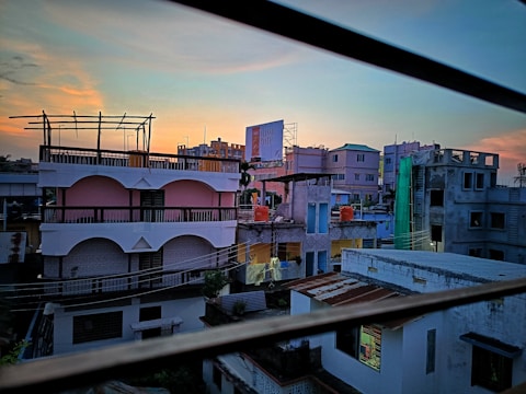 Urban setting with multiple residential buildings at sunrise or sunset, showcasing a mix of architectural styles. The sky has a gradient of blue and orange hues. There are utility lines in the foreground and a billboard in the background.