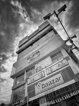 A multi-story commercial building featuring various shop advertisements, including those for clothing, is prominently shown against a dramatic cloudy sky. Large advertisements and store signage form distinct visual elements on the facade of the building. Streetlights and speaker poles appear in the foreground, creating an urban setting.