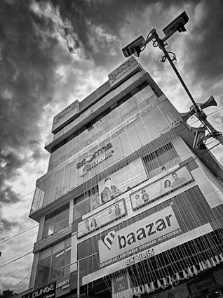 A multi-story commercial building featuring various shop advertisements, including those for clothing, is prominently shown against a dramatic cloudy sky. Large advertisements and store signage form distinct visual elements on the facade of the building. Streetlights and speaker poles appear in the foreground, creating an urban setting.