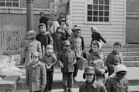 A group of children dressed in winter clothing are walking down the steps of a brick building. They appear cheerful and are accompanied by a woman in the background.