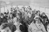 A group of young students sits inside a school bus, filling several rows of seats. The children are dressed in winter clothing, suggesting a cold season. The atmosphere inside the bus is lively, with many of them looking towards the camera.