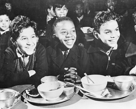 A group of children, smiling and seated at a table with empty bowls and cups, suggesting a school or communal meal setting. The focus is on three boys in the foreground, who appear to be enjoying each other's company.