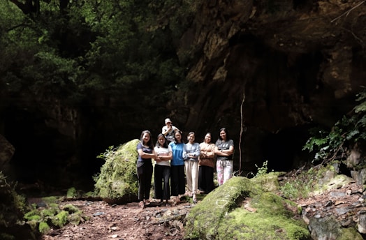 A group of people stand together in a lush, forested area with abundant greenery and moss-covered rocks. The setting appears to be a forest or cave entrance, with sunlight filtering through the trees.