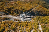 An aerial view of a mountainous landscape covered in dense, colorful forest with a mix of green and yellow foliage. A winding dirt road leads to a small industrial site with several buildings situated in a clearing among the trees.