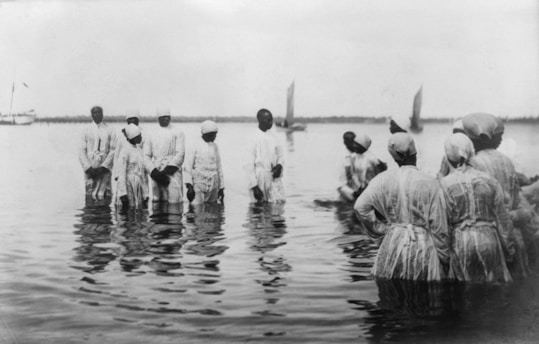 A group of individuals dressed in white robes standing in shallow water, possibly engaged in a ceremonial or religious activity. Sailboats float in the background, indicating a calm body of water.