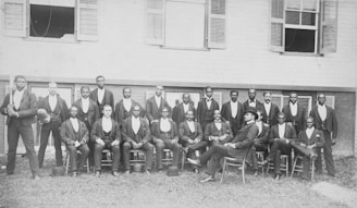 A group of men are posed in front of a building with several windows, wearing formal attire including jackets and bowties. The men are arranged in two rows, with those in the front row seated and those in the back row standing. Among them, one man is seated slightly apart wearing a different style of hat.