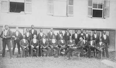 A group of men are posed in front of a building with several windows, wearing formal attire including jackets and bowties. The men are arranged in two rows, with those in the front row seated and those in the back row standing. Among them, one man is seated slightly apart wearing a different style of hat.