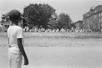 A young boy stands on a dirt path watching a large group of people gathered on a grassy slope across the road. The scene includes several buildings, utility poles, and a row of trees in the background.