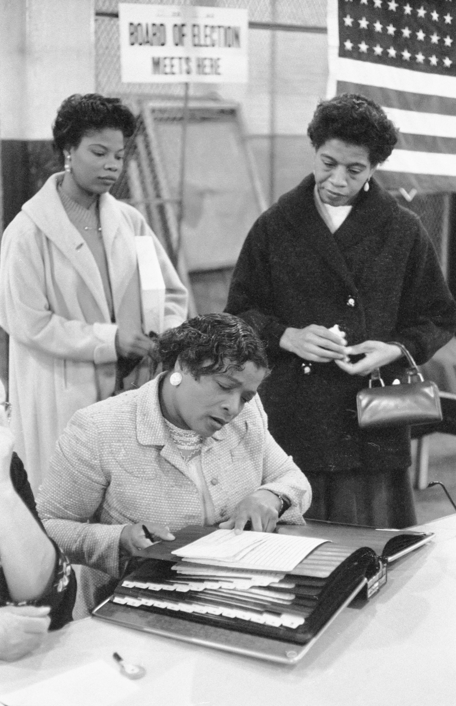 A black-and-white archival photograph of three Black women at a U.S. polling place in 1957, one examining a voter registration book.