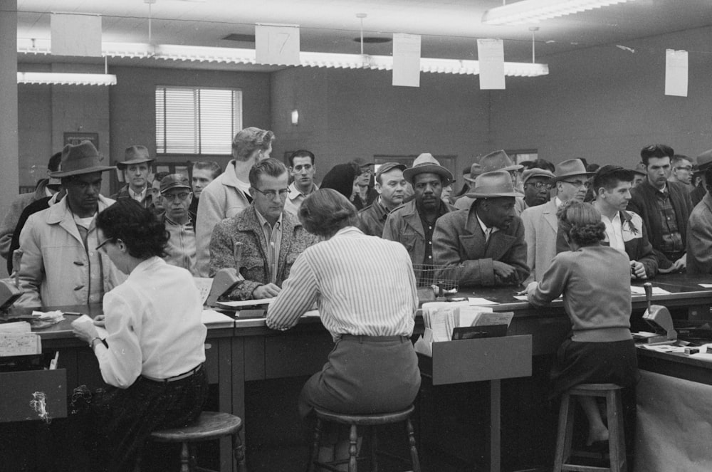 Unemployed men, including African Americans, line up at a counter for assistance from women office workers at an unemployment office, Detroit, Michigan.