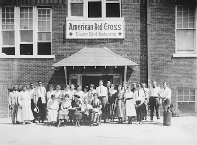 A group of men and women stand together in front of a brick building with a sign that reads 'American Red Cross Disaster Relief Headquarters.' The individuals are dressed in early 20th-century attire, with some seated while others stand in rows. The scene appears to be a formal gathering or a group photo of staff or volunteers.