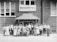 A group of men and women stand together in front of a brick building with a sign that reads 'American Red Cross Disaster Relief Headquarters.' The individuals are dressed in early 20th-century attire, with some seated while others stand in rows. The scene appears to be a formal gathering or a group photo of staff or volunteers.