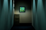 A dimly lit narrow hallway with smooth, light-colored walls. At the end of the hallway, a filing cabinet stands against the wall, partially illuminated. Above the cabinet, a small framed window displays vibrant green foliage bathed in sunlight, providing contrast to the interior's muted tones.
