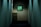 A dimly lit narrow hallway with smooth, light-colored walls. At the end of the hallway, a filing cabinet stands against the wall, partially illuminated. Above the cabinet, a small framed window displays vibrant green foliage bathed in sunlight, providing contrast to the interior's muted tones.