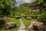 A lush garden landscape featuring a stone pathway leading through a grassy area surrounded by large rocks and dense green foliage. In the background, there is a multi-story building with a sloping roof and a thatched pavilion nestled among tall trees, with a mountainous backdrop against a cloudy sky.