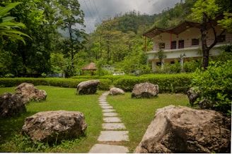 A lush garden landscape featuring a stone pathway leading through a grassy area surrounded by large rocks and dense green foliage. In the background, there is a multi-story building with a sloping roof and a thatched pavilion nestled among tall trees, with a mountainous backdrop against a cloudy sky.