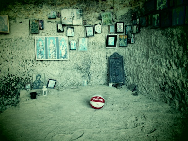 A rocky, cave-like room with numerous religious icons and framed images hanging on the walls. An ornate chair with intricate carvings is placed against the wall. In the foreground, a red and white basketball with the Spalding logo is prominently placed on the ground.