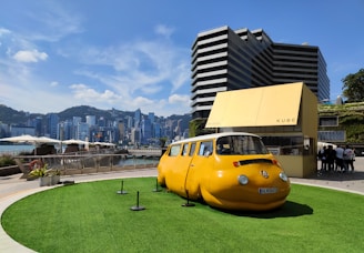 A unique yellow car with a round shape resembling a capsule is displayed on a grassy platform outdoors. The background features a modern cityscape with tall buildings and a mountainous horizon. There is a small modern structure labeled 'KUBE' nearby, and a group of people is gathered around it. The sky is clear with a few clouds.