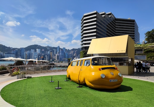 A unique yellow car with a round shape resembling a capsule is displayed on a grassy platform outdoors. The background features a modern cityscape with tall buildings and a mountainous horizon. There is a small modern structure labeled 'KUBE' nearby, and a group of people is gathered around it. The sky is clear with a few clouds.