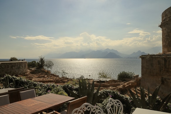 A picturesque coastal view with a serene seascape, featuring calm waters reflecting the sunlight and a distant mountain range under a partly cloudy sky. In the foreground, there is a terrace with wooden tables and ornate chairs, surrounded by lush green plants and stone structures.