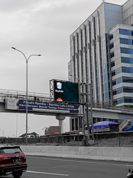 An urban scene featuring a multi-lane highway with vehicles including a red SUV and a large truck. Above the road, an overpass with a street sign in a foreign language is visible. A digital billboard displaying 'Recycle Bin' is mounted on the overpass. Large modern office buildings with reflective glass windows dominate the background.