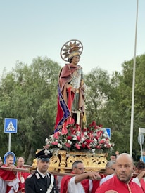 A religious procession features a large statue of a saint adorned with ornate clothing and a halo, mounted on a platform decorated with red and white flowers. Several individuals, some in uniform and others in red and white robes, are carrying the platform. Trees are visible in the background along with several street signs.