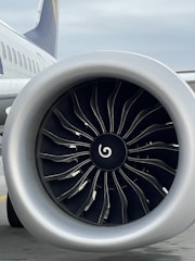A close-up view of a modern airplane jet engine, showing its metallic fan blades and smooth circular structure. The aircraft's body and part of the wing are visible in the background.