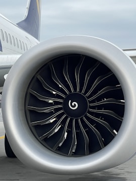 A close-up view of a modern airplane jet engine, showing its metallic fan blades and smooth circular structure. The aircraft's body and part of the wing are visible in the background.