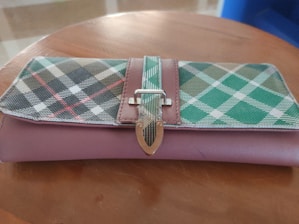 A plaid-patterned wallet with diagonal stripes in green, black, and red on top of a pink surface. The wallet features a metallic buckle clasp and is resting on a wooden table.