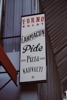 A vertical rectangular sign featuring text in both red and black colors. The sign promotes various food items including Lahmacun, Pide, Pizza, and Kahvalti. It is affixed to a building with a corrugated metal exterior and has an orange-red awning partially visible on the left side.