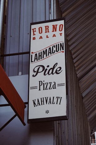 A vertical rectangular sign featuring text in both red and black colors. The sign promotes various food items including Lahmacun, Pide, Pizza, and Kahvalti. It is affixed to a building with a corrugated metal exterior and has an orange-red awning partially visible on the left side.