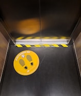 An elevator interior featuring a yellow and black caution tape at the door threshold and an orange floor sticker with footprints and French text about maintaining distance.