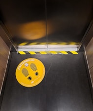 An elevator interior featuring a yellow and black caution tape at the door threshold and an orange floor sticker with footprints and French text about maintaining distance.