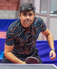 A person is focused and leaning towards a ping pong table, holding a paddle in a ready position. The individual is wearing a patterned shirt with the brand name repeated across it and a wristwatch on their right wrist. The background shows a table tennis setting with barriers and walls.