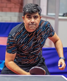 A person is focused and leaning towards a ping pong table, holding a paddle in a ready position. The individual is wearing a patterned shirt with the brand name repeated across it and a wristwatch on their right wrist. The background shows a table tennis setting with barriers and walls.