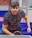 A person is focused and leaning towards a ping pong table, holding a paddle in a ready position. The individual is wearing a patterned shirt with the brand name repeated across it and a wristwatch on their right wrist. The background shows a table tennis setting with barriers and walls.