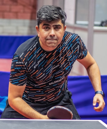 A person is focused and leaning towards a ping pong table, holding a paddle in a ready position. The individual is wearing a patterned shirt with the brand name repeated across it and a wristwatch on their right wrist. The background shows a table tennis setting with barriers and walls.