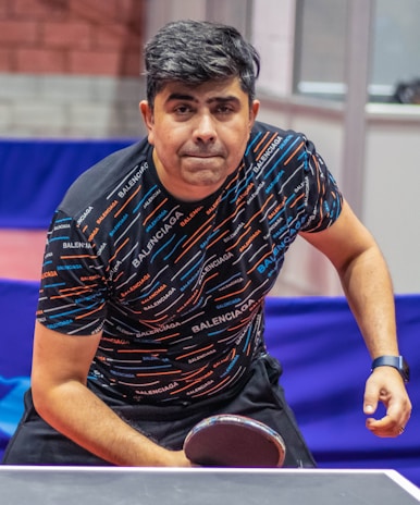 A person is focused and leaning towards a ping pong table, holding a paddle in a ready position. The individual is wearing a patterned shirt with the brand name repeated across it and a wristwatch on their right wrist. The background shows a table tennis setting with barriers and walls.